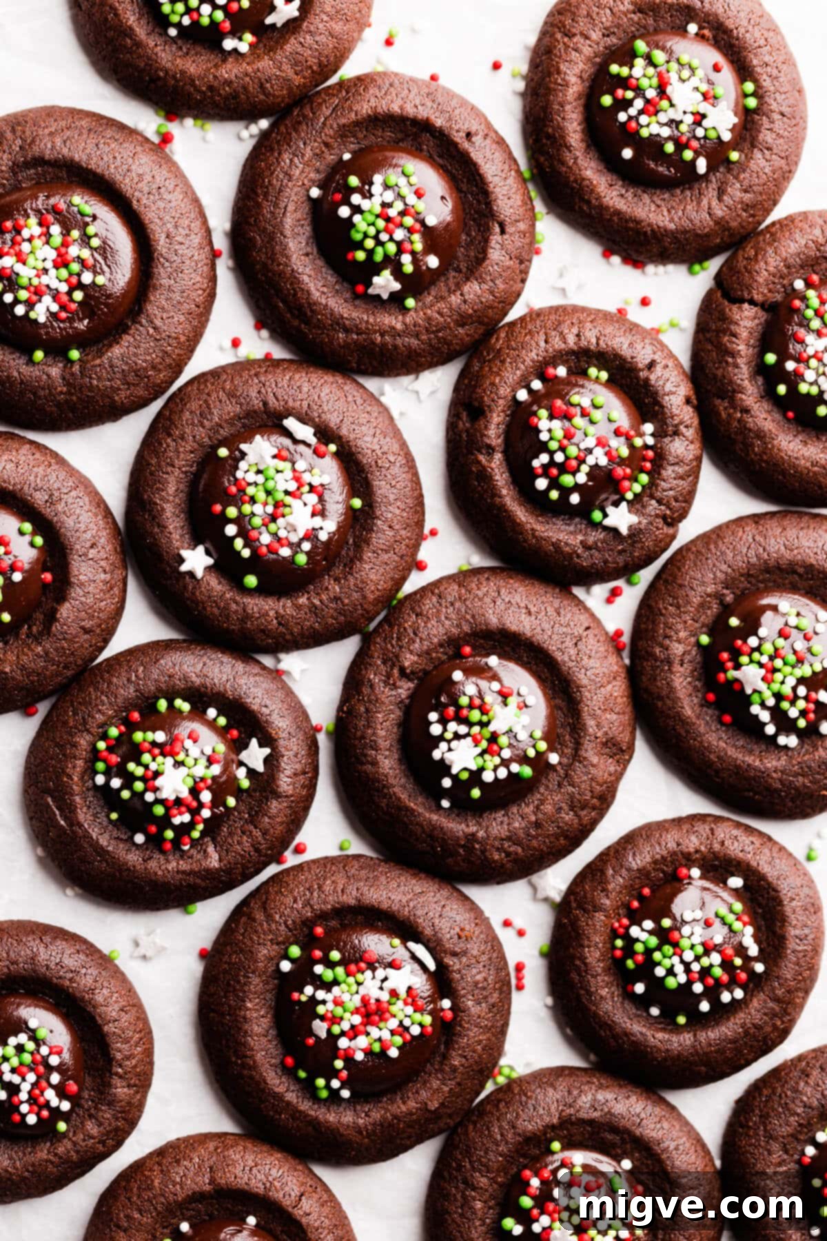 Top view close-up of deeply rich chocolate thumbprint cookies with shiny ganache and colorful sprinkles on a white surface, ready to be enjoyed.